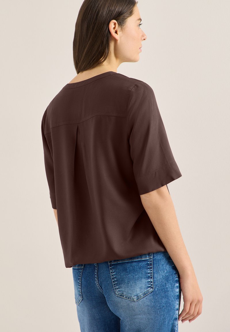 Femme portant un haut ample à manches courtes de couleur marron foncé et un jean en denim bleu, vue de dos sur un fond beige uni.