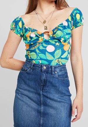 Frau trägt ein blaues, florales schulterfreies Oberteil mit einem vorderen Ringdetail und einen blauen Jeansrock, kombiniert mit mehrlagigen Halsketten.