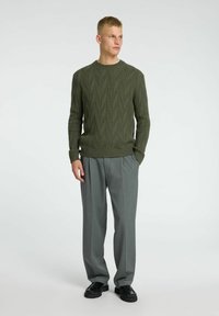 Alyvuogių spalvos megztinis su lapų raštu, derinamas su pilkais klostuotais kelnėmis ir juodais batais. Lygus audinys, raukšlėtos rankogaliai ir apačia.