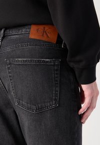 Les jeans en denim noir présentent une texture délavée, des coutures détaillées et un patch de taille en cuir marron avec "Calvin Klein" en relief.