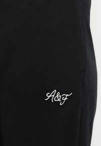Črne trenirke iz mehke tkanine z belim vezenim logotipom "A&F" na spodnji levi strani. Enostavna zasnova s čistimi linijami.