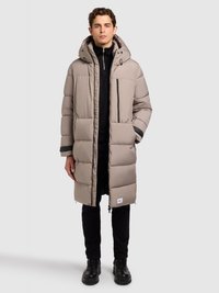 Svijetlo bež puhasti kaput s kapuljačom, patentnim zatvaračem sprijeda, bočnim džepovima i crnim detaljima. Nošen preko crne majice i crnih hlača, upotpunjen crnim čizmama.