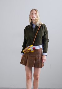 Femeie blondă îmbrăcată cu un pulover cu fermoar de culoare verde închis, o cămașă albastră, o fustă plisată maro și o geantă tip crossbody în formă de o pasăre colorată.