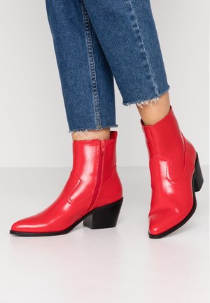 Kobieta w niebieskich, krótko przyciętych jeansach i błyszczących czerwonych botkach z czarnymi, masywnymi obcasami i bocznymi zamkami, stojąca na białej podłodze.