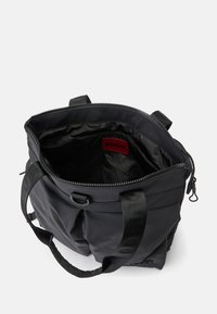 Sac fourre-tout noir avec ouverture à zip, deux poignées et intérieur montrant une étiquette rouge HUGO sur une doublure noire.