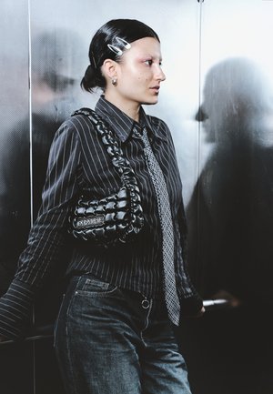 Young woman with dark hair, wearing black pinstripe shirt, patterned tie, and jeans, standing in an elevator holding the handrail.
