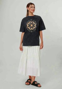 Ženska, oblečena v črno majico z grafiko sonca in zvezd, belo večplastno krilo, črne sandale z zaponkami, stoji pred enobarvno svetlo ozadje.