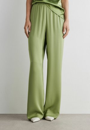 Person trägt lockere, hellgrüne Satin-Hose und passendes Oberteil, steht vor einem einfarbigen hellen Wand- und Bodenhintergrund.