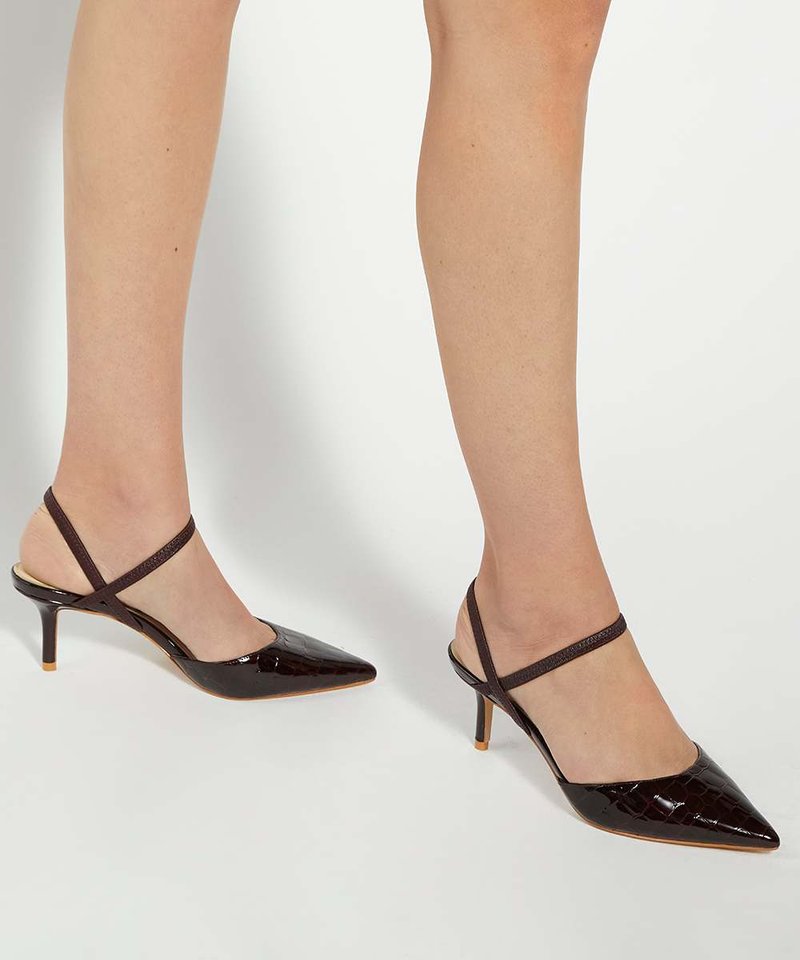 Benen met donkerbruine puntige kittenheels met dunne kruislings gespen aan de enkel op een effen witte achtergrond.