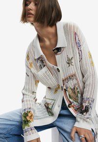 Balta megzta kardiganas su gėlių raštais, turintis apykaklę, mygtukų užsegimą ir laisvą siluetą. Medžiaga yra pralaidi orui ir lengva.