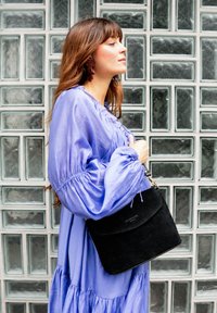 Frau in einem lavendelfarbenen Kleid mit Puffärmeln hält eine schwarze Schultertasche und steht seitlich vor einer Wand aus strukturierten Glasbausteinen.