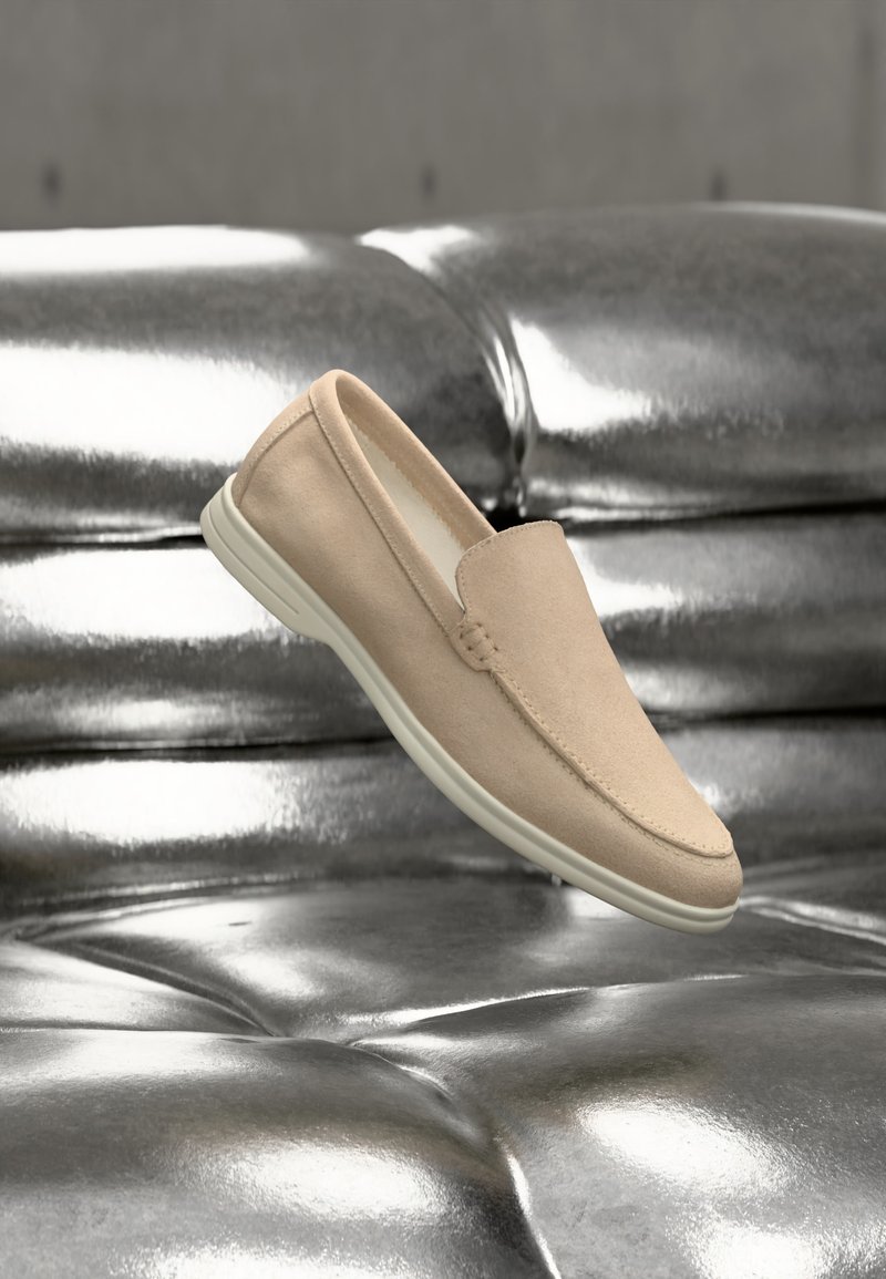 Μπεζ σουέντ παπούτσι slip-on με λευκή ελαστική σόλα, αιωρούμενο πάνω από επιφάνεια από γυαλιστερό ασημί δερμάτινο καπιτονέ.
