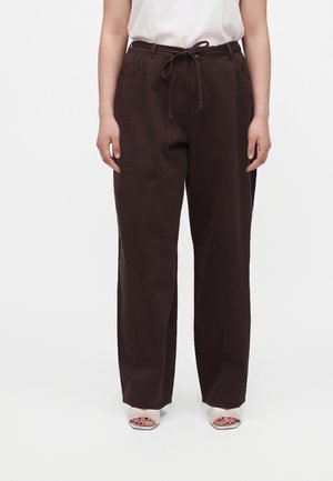 Femme portant un pantalon ample marron foncé avec une taille à cordon, un haut blanc sans manches et des chaussures blanches à bouts ouverts.