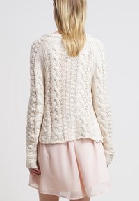 Elfenbensfärgad kabelstickad tröja med en texturerad design, med långa ärmar och en något kortad baksida, tillsammans med en genomskinlig rosa klänning.