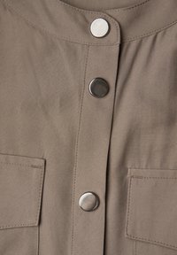 Khakifarbenes Hemd mit rundem Ausschnitt, ausgestattet mit drei silbernen Druckknöpfen und zwei Vorderentaschen mit glatter, matter Textur.