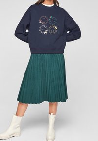 Marinblå sweatshirt med lekfullt leende ansikte-tryck, i kombination med en plisserad turkos kjol och vita ankelstövlar. Outfiten kombinerar avslappnade och stiliga element.