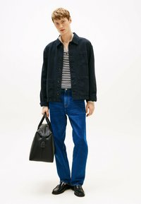Marineblauwe katoenen jas over een gestreept shirt, blauwe denim jeans, zwarte leren tas en zwarte schoenen. De outfit heeft een casual ontwerp met zakken.