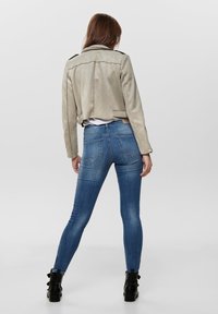 Beige suède cropped jack met zwarte accenten, gecombineerd met blauwe skinny jeans en zwarte enkellaarsjes met een strikdetail aan de achterkant.