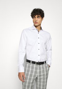 Camicia bianca a maniche lunghe con vestibilità slim, bottoni neri e colletto a contrasto, abbinata a pantaloni grigi a quadri e una cintura nera.