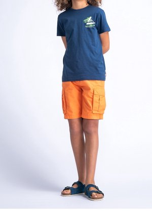 T-shirt blu in cotone con stampa grafica verde, abbinata a pantaloni cargo arancioni e sandali blu con dettaglio a fibbia.