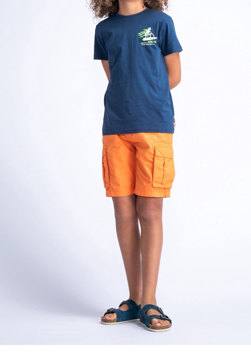T-shirt en coton bleu avec imprimé graphique vert, associé à un short cargo orange et des sandales bleues avec une boucle.