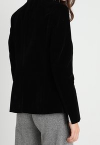 Černý sametový blejzr s přiléhavým střihem, dlouhými rukávy a klopovým límcem. Obsahuje hladkou texturu a diskrétní zadní rozparek pro pohodlný pohyb.