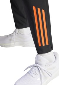 Schwarze Sporthosen mit drei vertikalen orangen Streifen an der Unterschenkel. Kombiniert mit weißen Sportschuhen und weißen Socken.
