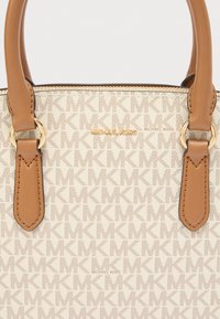 Μπεζ υφασμάτινη τσάντα ώμου με ανάγλυφο σχέδιο λογότυπου, καφέ δερμάτινες λαβές και μεταλλικά εξαρτήματα σε χρυσή απόχρωση. Διαθέτει κλείσιμο με φερμουάρ και λεπτομέρειες με το όνομα της μάρκας.