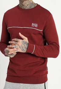 Sudadera burdeos con franja de acento blanca y logo bordado "BLOCK". Puños y dobladillo acanalados, textura suave y diseño clásico de cuello redondo.