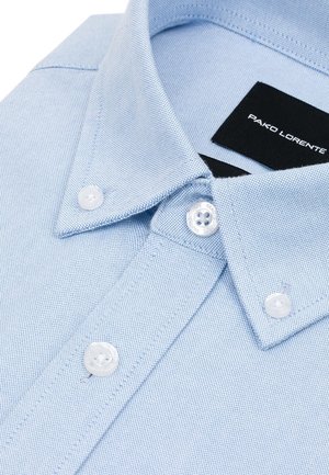 Jasnoniebieska koszula zapinana na guziki z teksturowanej tkaniny, z spiczastym kołnierzykiem i białymi guzikami. Na kołnierzyku widoczna jest metka "PAKO LORENTE".