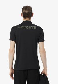 Polo negro de algodón con cuello y mangas cortas. Presenta un gran texto verde "LACOSTE" en la espalda. Textura lisa, diseño casual.