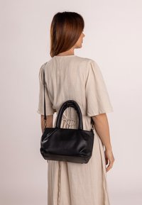 Czarna skórzana torba o luźnym fasonie, z plecioną rączką i odczepianym paskiem na ramię, wyeksponowana na neutralnym tle.