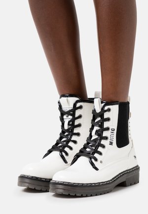 Hvide læderankelstøvler med snørebånd, sorte såler, sorte snørebånd, sorte elastikpaneler og "MUSTANG" branding, båret på mørkehudede ben.