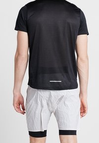 Černé krátké sportovní tričko s hladkou texturou, spárované s bílými zmuchlanými sportovními šortkami a černými kompresními šortkami pod nimi.