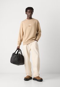 Beige sweatshirt with a small logo patch, white wide-leg trousers, black bag with handles, and brown slip-on shoes with textured soles.