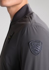 Gray bomber jacket featuring a flat collar, a zippered front, and a textured embroidered logo patch on the left shoulder.