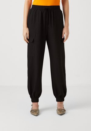 Persoon draagt zwarte cargobroek met elastische taille en omgeslagen enkels, en gouden slingback flats met puntige neus, staand tegen een effen achtergrond.