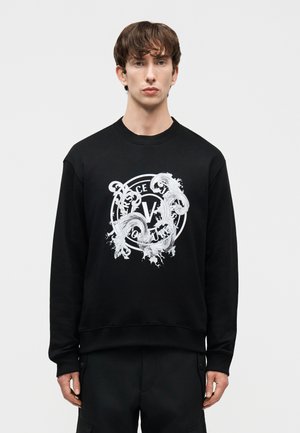 Jaunas vyras dėvi juodą džemperį su balto ornamentuoto apskrito „Versace Jeans Couture“ logotipo spauda, stovintis prie paprasto šviesaus fono.
