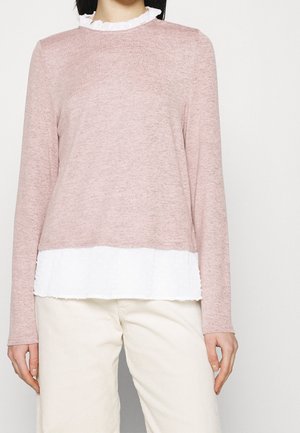 Kvinde iført lyserød langærmet top lagdelt over en hvid flæset skjorte, kombineret med off-white bukser mod en ensfarvet baggrund.