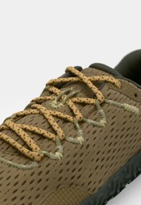 Zapato deportivo marrón perforado con cordones verdes y amarillos, parte superior texturizada, punta reforzada y suela de goma con patrones de tracción.