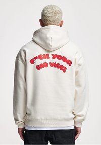 Maglione con cappuccio color crema dotato di una grande grafica in lettere bubble rossa sul retro con la scritta "F*CK YOUR BAD VIBES". Realizzato in tessuto morbido e con vestibilità rilassata.
