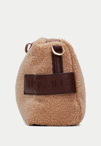 Sac en tissu peluche beige, de forme arrondie, avec une bandoulière en cuir brun foncé et des détails en métal doré. Extérieur texturé et doux.