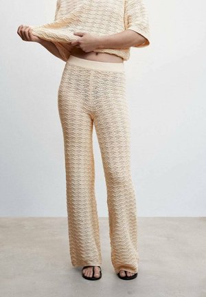 Personne portant un pantalon beige en tricot et un haut assorti, levant légèrement le haut pour montrer le ventre, debout sur un sol uni.