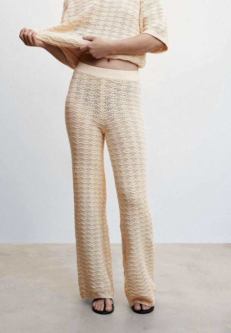 Personne portant un pantalon beige en tricot et un haut assorti, levant légèrement le haut pour montrer le ventre, debout sur un sol uni.