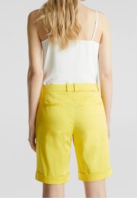 Pantalones cortos bermuda amarillos con textura suave, dobladillo enrollado y trabillas para cinturón, combinados con una camiseta sin mangas blanca. Vista trasera.