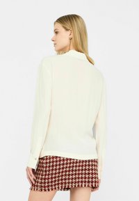 Camicia cremosa a maniche lunghe, con polsini abbottonati; abbinata a una gonna con fantasia houndstooth rossa, con frange all'orlo.