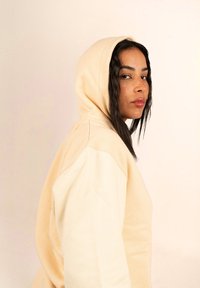 Sudadera con capucha de color beige claro y textura suave, con un corte holgado y costuras laterales, modelada por una persona con cabello largo y oscuro.
