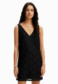 Čierne čipkové šaty s výstrihom do V, bez rukávov, so vzormi kvetov a textúrovaným povrchom. Doplnky v striebornom tóne s náhrdelníkom a náramkom v tvare srdca.