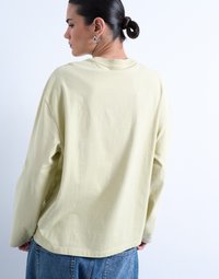 Γυναίκα με σκούρα μαλλιά σε κοτσίδα χαμηλά, φορούν ασημένια σκουλαρίκια, μπλούζα μακρυμάνικη σε απαλό κίτρινο χρώμα και μπλε τζιν, γυρισμένη από την πλάτη, πάνω σε απλό φόντο.