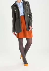 Grijze wollen jas met gouden knopen, lichtblauw overhemd, oranje rok, grijze panty’s en gele hoge hakken. Eenvoudig ontwerp en getailleerde pasvorm.